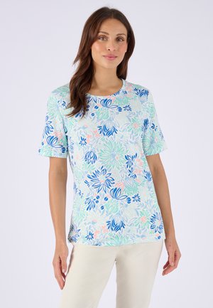 Vrouw die een kort mouws bloemen top draagt met blauwe, groene en roze bladeren en bloemen, gecombineerd met lichtbeige broek, staand tegen een witte achtergrond.