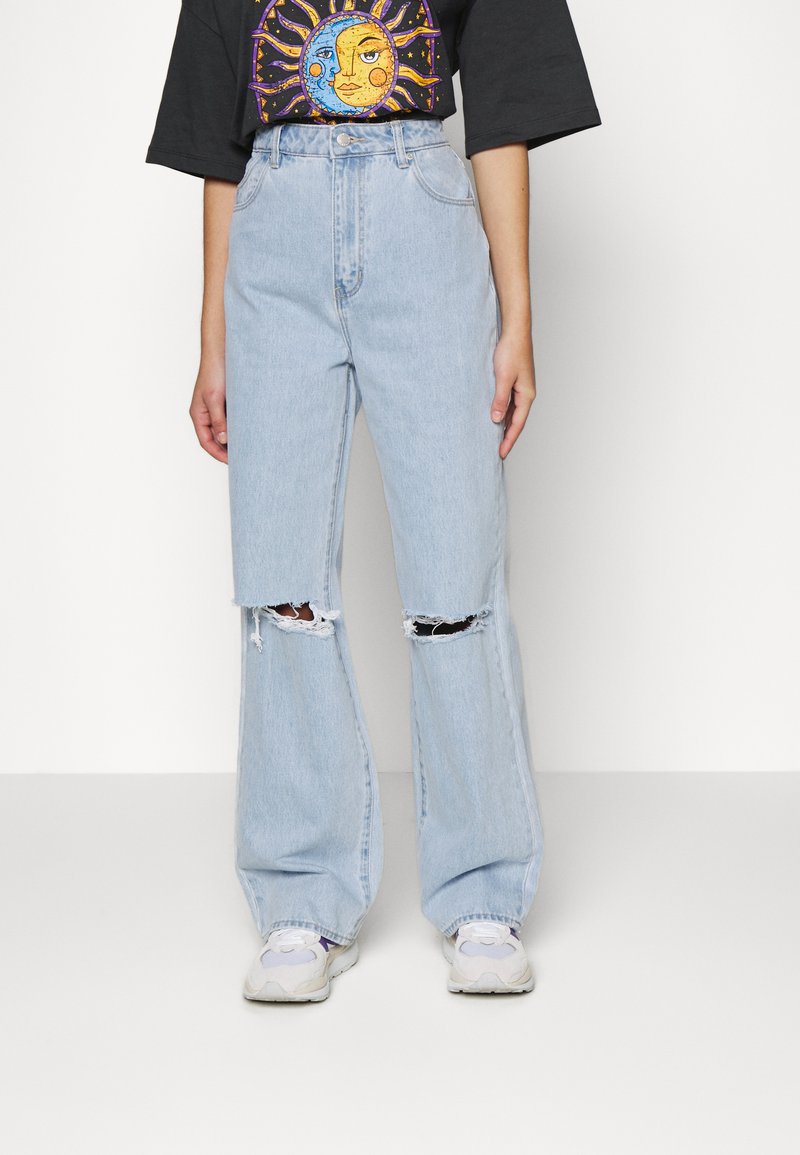 Jeans de ganga azuis claros de perna larga com recortes nos joelhos, de cintura alta e bainha desfiada. Combinados com uma t-shirt gráfica preta e sapatilhas.