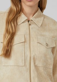 Beżowa kurtka z korduroyu z pełnym zamkiem błyskawicznym, spiczastym kołnierzem i dwiema przednimi kieszeniami. Teksturowany materiał z subtelnymi pionowymi liniami.