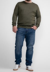 Man wearing olive green sweater, blue jeans, and white sneakers standing against a plain white background with one hand in pocket.