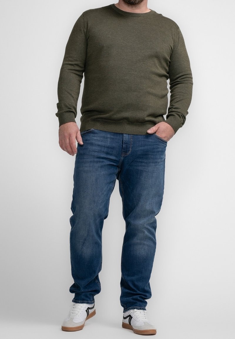 Man wearing olive green sweater, blue jeans, and white sneakers standing against a plain white background with one hand in pocket.