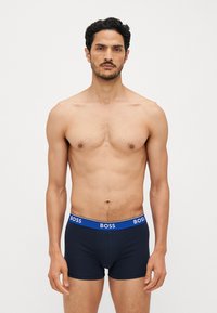 Boxeri navy cu talie albastră, având inscripția "BOSS" în text alb; material elastic, croială strânsă și design simplu. Modelul stă în picioare.