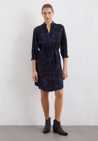 Robe bleu marine avec des motifs abstraits, fermeture à boutons sur le devant, manches trois-quarts et ceinture à la taille. Associée à des bottines noires.