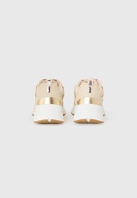 Sneaker beige con accenti metallici dorati, superiore testurizzato e suola bianca. Presenta una striscia riflettente sul tallone e un colletto imbottito.