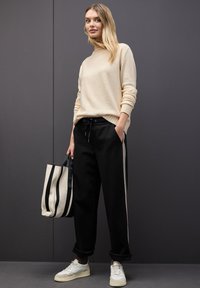 Beige tröja, svarta sweatpants med sidostrikar och en randig totebag. Skorna är vita med en beige accent. Släta texturer genomgående.