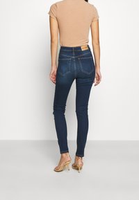 Mujer de pie, vestida con jeans ajustados azul oscuro y una blusa de manga corta beige, combinada con sandalias de tacón alto beige, contra un fondo liso.