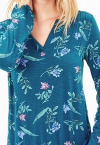 Långärmad teal topp med blommönster i rosa och blått, med en V-halsringning och subtil textur. Tyget verkar mjukt och lätt.