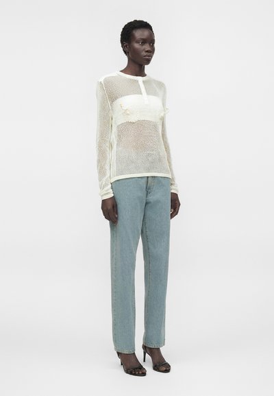 Femme debout portant un haut en maille blanche à manches longues sur un bandeau blanc, un jean bleu clair et des sandales noires à talons ouverts.