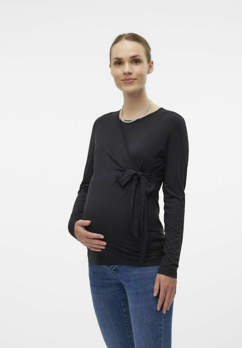 Top premaman nero a maniche lunghe con dettaglio del nodo laterale. Tessuto morbido, design aderente, indossato con jeans in denim blu, finitura liscia.