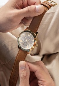 Orologio con cinturino in pelle marrone, cassa dorata, quadrante argentato, grandi numeri e tre sottodial di cronografo.