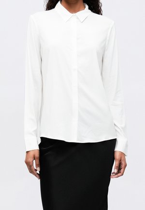 Femme portant une chemise blanche à manches longues avec un col pointu, associée à une jupe noire, debout devant un fond uni.