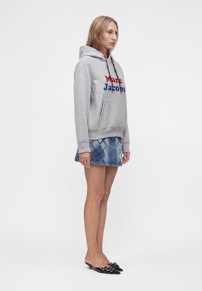 Γυναίκα που φοράει γκρι φούτερ Marc Jacobs, φούστα denim patchwork και μαύρα μυτερά παπούτσια mule, στέκεται πλαγίως σε λευκό φόντο.