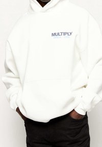 Bílá mikina s kapucí s nápisem "MULTIPLY DREAMERS" v modré a tmavě modré barvě. Obsahuje přední kapsu a žebrované manžety. Textura působí měkce.