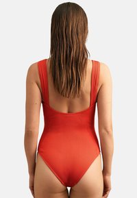 Maillot de bain une pièce rouge à côtes avec des bretelles larges et un dos décolleté. Texture lisse avec une ouverture de jambe haute.