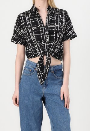 Vrouw draagt een zwart-wit geruit kortarm blouse met strik aan de voorkant, gecombineerd met een high-waist blauwe spijkerbroek, staand tegen een effen achtergrond.