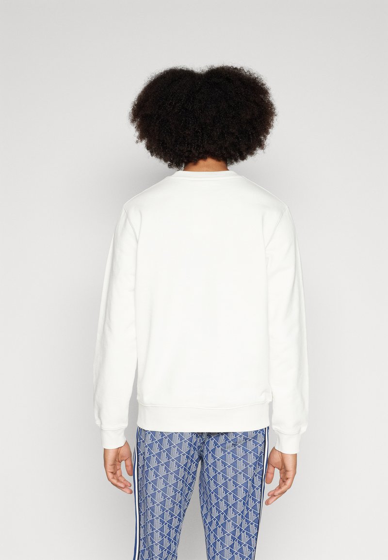 Felpa bianca con collo rotondo e polsini a coste, indossata sopra leggings blu a fantasia con un motivo a diamante e strisce laterali.