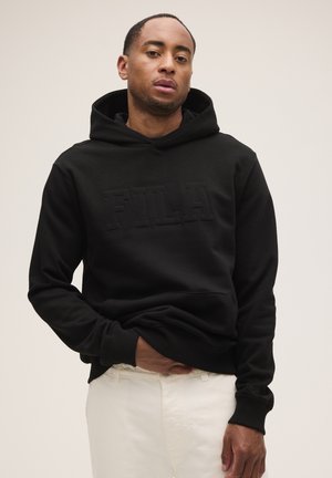 Un hombre luce una sudadera negra con capucha de FILA y unos pantalones blancos sobre un fondo liso y claro.
