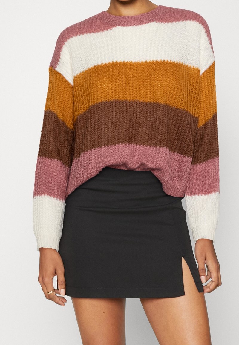 Un maglione multicolore presenta righe orizzontali in rosa, bianco, senape, marrone e crema, con una vestibilità ampia e una texture a costine. Abbinato a una minigonna nera.