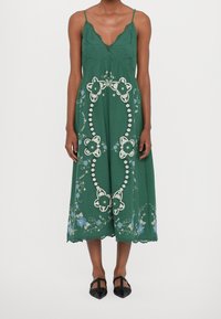 Zelené bezrukávové šaty s květinovou výšivkou, krajkovým lemem, V výstřihem a volným A-liniovým tvarem. Nošeny s černými špičatými botami.
