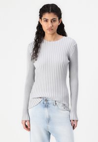 Lys grå ribbestrikket langermet topp med rund hals, har en slim fit og sidesplitter i kanten. Kombinert med lyseblå bukser.