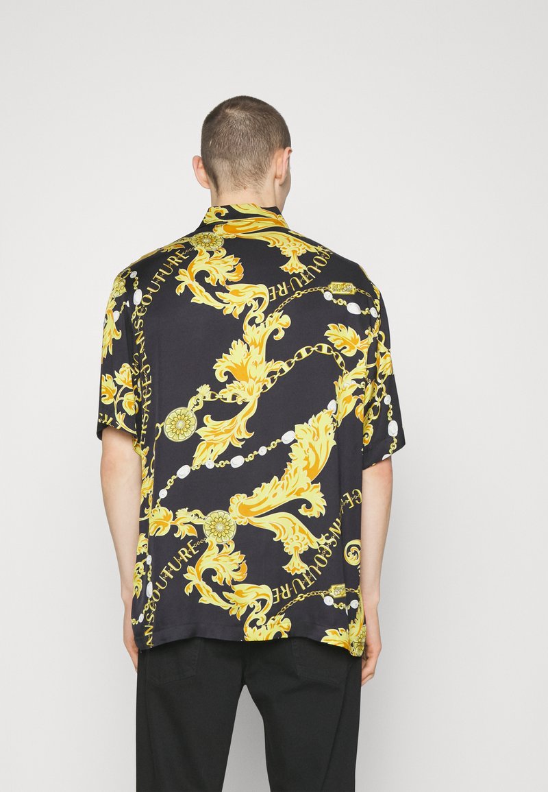 Chemise à manches courtes en tissu noir avec des motifs baroques jaunes audacieux, comprenant des chaînes et des accents décoratifs. Coupe décontractée avec un col.