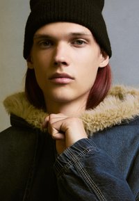 Jeansjacka med en pälsfodrad huva, i blå färg, ribbad svart beanie och en mjuk textur. Händerna greppar kragen.