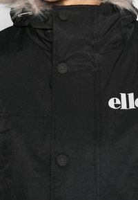 Parka de invierno negra con capucha con borde de piel, presenta múltiples botones negros y un logo de la marca impreso en blanco en la parte delantera. Textura suave.