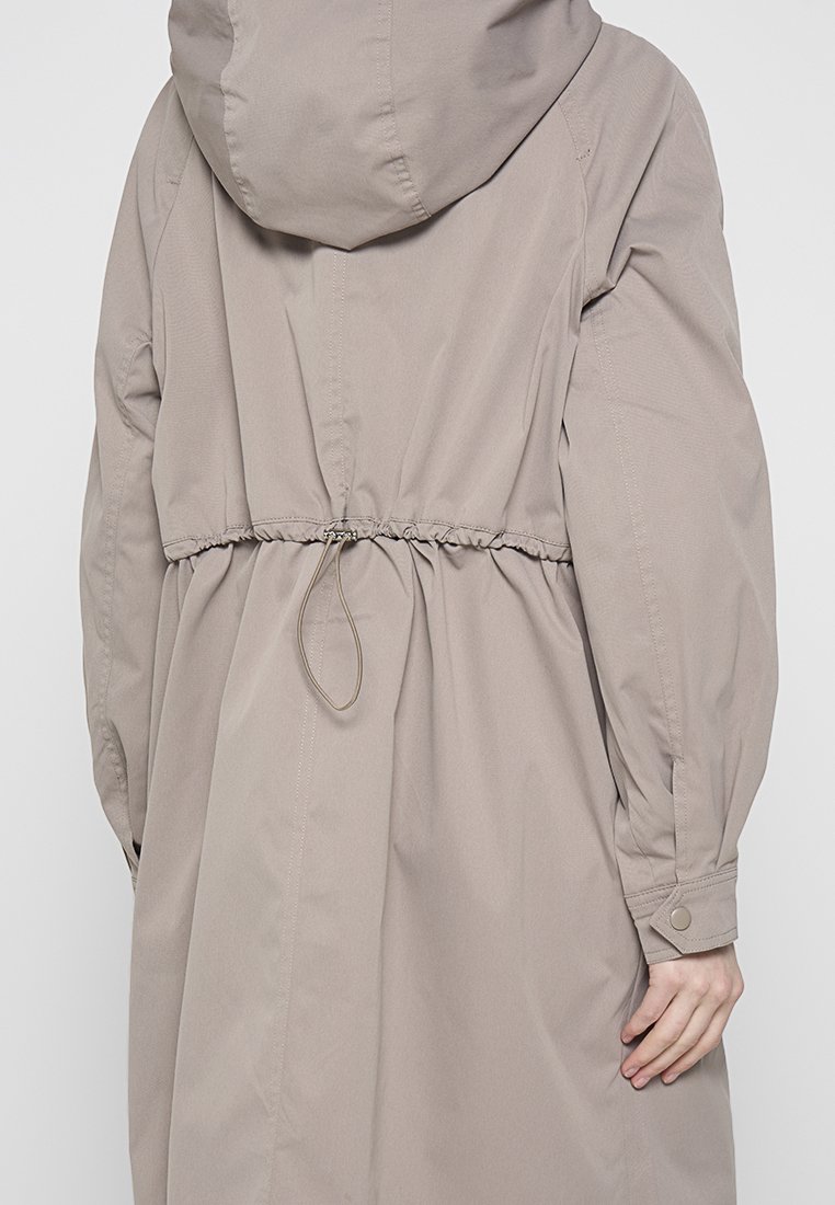 Manteau capuche taupe clair avec taille à cordon, manches longues et poignets boutonnés. Tissu lisse et design décontracté.