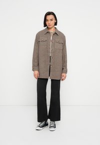 Καρό πουκάμισο-ζακέτα σε καφέ και μαύρο, με χαλαρή εφαρμογή, δύο τσέπες στο στήθος και κουμπώματα με κουμπιά. Συνδυάζεται με μαύρο παντελόνι καμπάνα και αθλητικά παπούτσια.