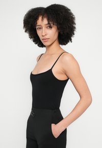 Melnas pieguļošas topiņš ar plānām lencēm, gludu tekstūru un zemu izgriezumu; kombinēts ar melnām augsta vidukļa biksēm ar apaļu aparatūras detaļu.