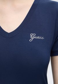Μπλε ναυτικό T-shirt με V-λαιμόκοψη και κοντά μανίκια με λογότυπο "Guess" από μικρές λευκές στρας στο αριστερό στήθος.