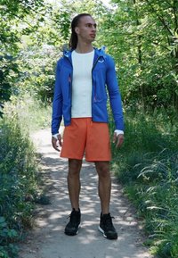 Sportstøj bestående af en blå zip-up jakke med orange detaljer, en tætsiddende hvid top og orange shorts; båret med sorte sneakers.