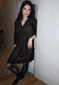 Dunkelbraunes, langärmliges Hemdkleid mit geknöpftem Verschluss, Rüschenkragen und gestuftem Design, kombiniert mit schwarzen Stiefeletten und durchsichtigen schwarzen Strumpfhosen.