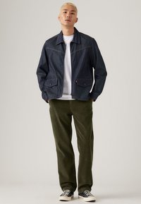 Dunkelblaue Jeansjacke mit zwei Fronttaschen, weißem Hemd darunter, olivgrünen Cordhosen und schwarzen Sneakers mit weißen Sohlen.