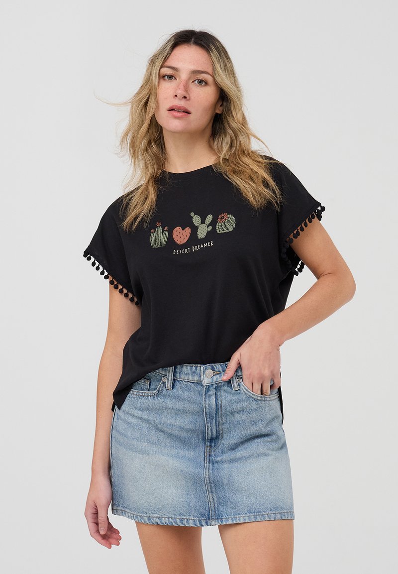 T-shirt nera con grafica di un cactus e il testo "DESERT DREAMER". Presenta un bordo in pom-pom sulle maniche. Abbinato a una mini gonna di denim chiaro.