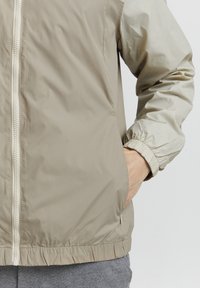 Lätt beige jacka med elastiska ärmmuddar, en framsida med dragkedja och sidofickor, tillverkad av ett jämnt, vattenavvisande material.