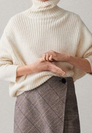 Person trägt einen cremefarbenen, gerippten Rollkragenpullover und einen grau-karierten Rock, die Hände locker vor dem Körper verschränkt.
