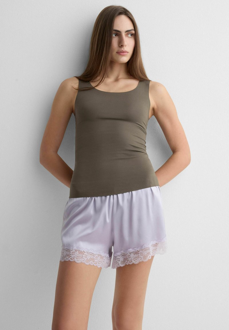 Mujer con camiseta sin mangas de color oliva y pantalones cortos de satén púrpura claro con borde de encaje, de pie contra una pared blanca lisa, mirando hacia un lado.