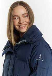 Jachetă căptușită de culoare bleumarin, cu guler înalt și fermoar. Prezintă broderie cu logo și un design de ancoră pe mânecă. Textură netedă.