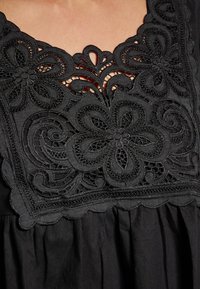 Top din țesătură neagră cu broderie florală intricată, guler cu margine ondulată și detalii decupate, evidențiind textura și designul.