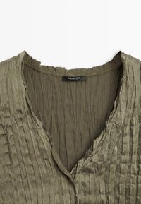 Camicia verde oliva con trama, scollo a V e bordi sfrangiati; presenta pieghe verticali e un'etichetta con la scritta "Massimo Dutti".