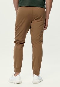 Brūni sporta bikses ar sašaurinātiem galiem, elastīgām aprocēm un gludu tekstūru. Jostasvieta ar smalku šuvju detaļu. Nēsātas ar baltām kedām.