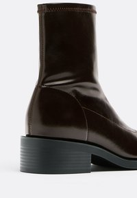 Bottine en cuir marron avec une finition brillante, dotée d'un talon block et d'une couture contrastante le long des coutures, offrant une silhouette élégante.