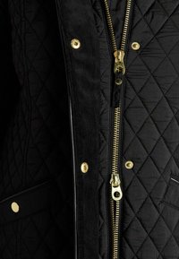 Melnā quiltētā jakā ar zelta rāvējslēdzēju, dimanta rakstu un zelta pogām. Strukturēta auduma ar gludām akcentiem.