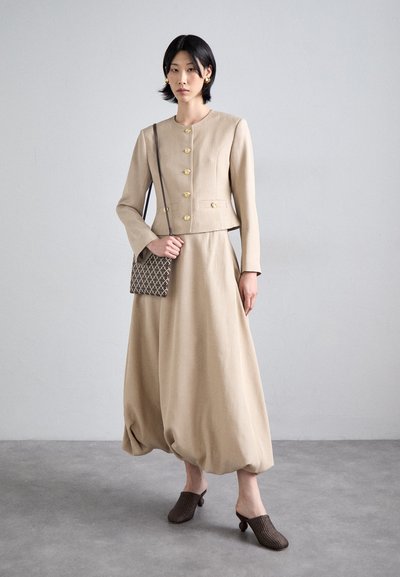 Veste en lin beige avec des boutons dorés et une jupe volumineuse assortie ; associé à des mules brunes texturées et un sac à bandoulière à motifs.