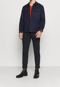 Tmavě modrá košile s límečkem přes červené tričko s výstřihem do V, doplněná tmavými džíny a černými chelsea botami. Obsahuje dvě náprsní kapsy.