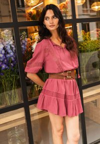 Vestido rosa de corte relajado, con detalles de encaje y falda acampanada. Acompañado de un cinturón marrón con una hebilla rectangular.