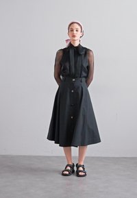 Black button-down dress with sheer sleeves and a flared skirt. Model wears a pink headscarf and black flat sandals, against a light background.
