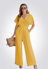 Une combinaison jaune à jambes larges avec un décolleté en V, des manches courtes et un tissu texturé. Associée à une pochette marron à franges et des lunettes de soleil.