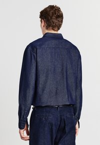 Camicia in denim scuro con maniche lunghe, polsini a bottoni e colletto classico. La texture è liscia con una sottile lucentezza; vestibilità su misura.